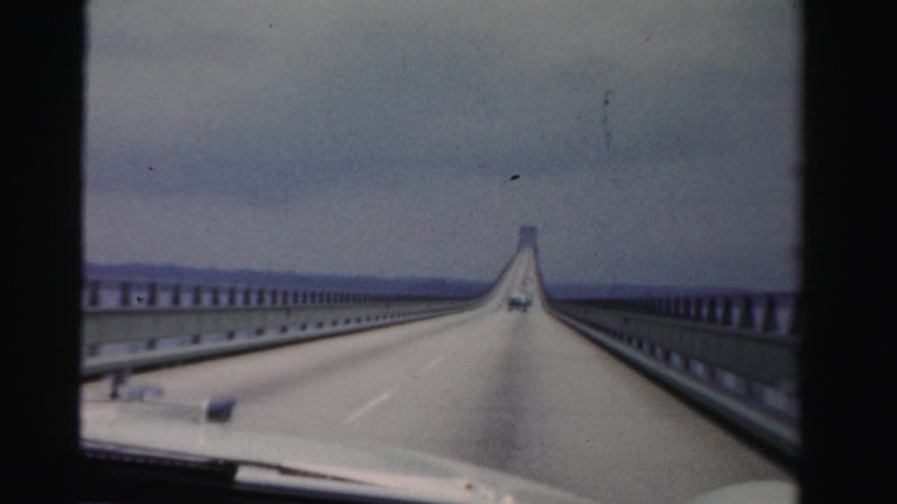 View from a car on a long causeway with guardrails, water, overcast sky, and a vehicle ahead.