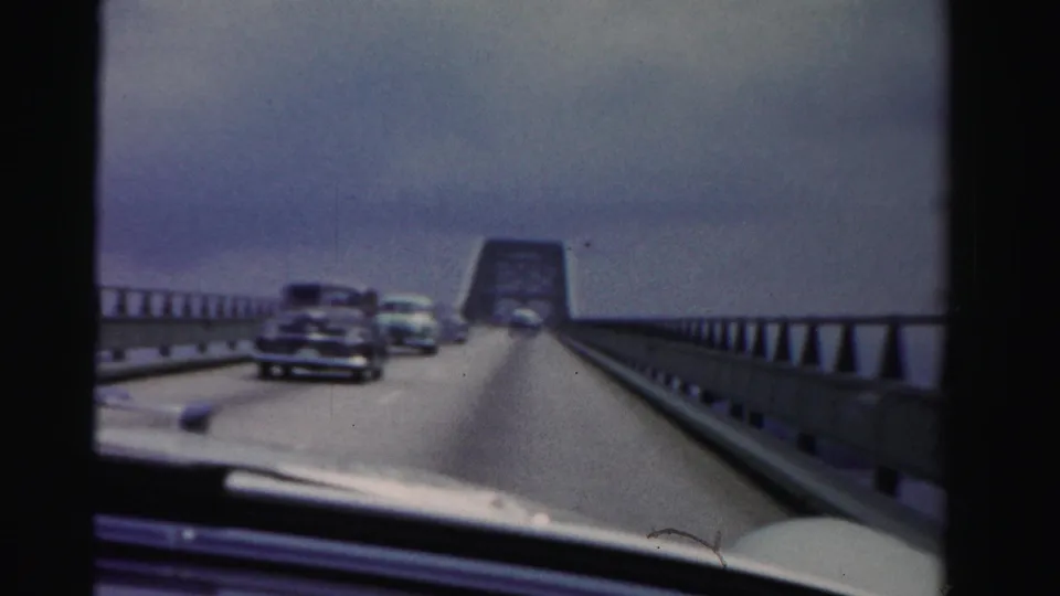 1955 Dade County 8mm frame showing oncoming mid-century cars on a long bridge, filmed through a windshield under gray sky.
