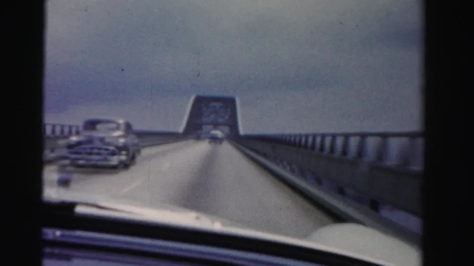 Close windshield view from 1955 Dade County footage as a rounded mid-century car passes on the bridge near the raised span.