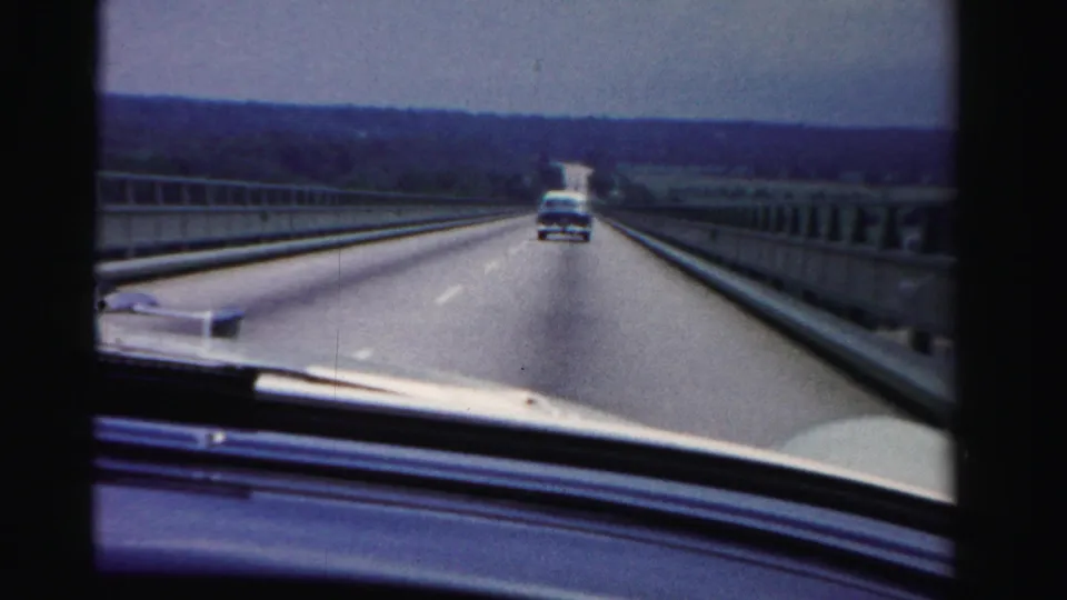 1955 Dade County car-window perspective showing a highway lane, roadside barriers, dashboard edge, and a mid-century vehicle ahead.
