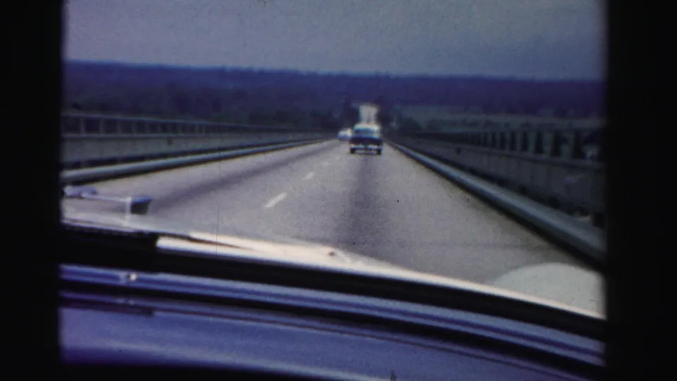 1955 Dade County dashboard view from a moving automobile, with a divided roadway, guardrails, distant trees, and traffic ahead.