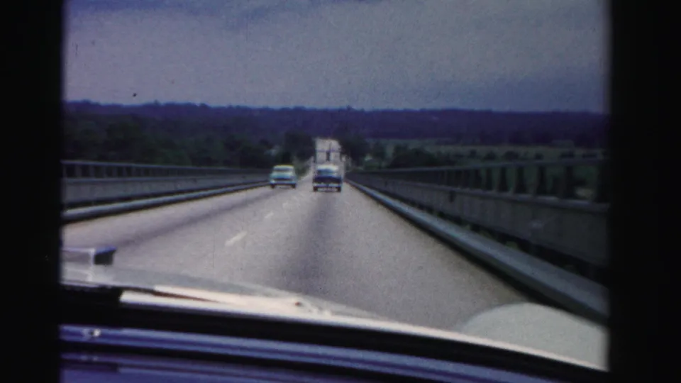 Windshield view on a Dade County bridge in 1955, with period cars traveling ahead and trees across the horizon.