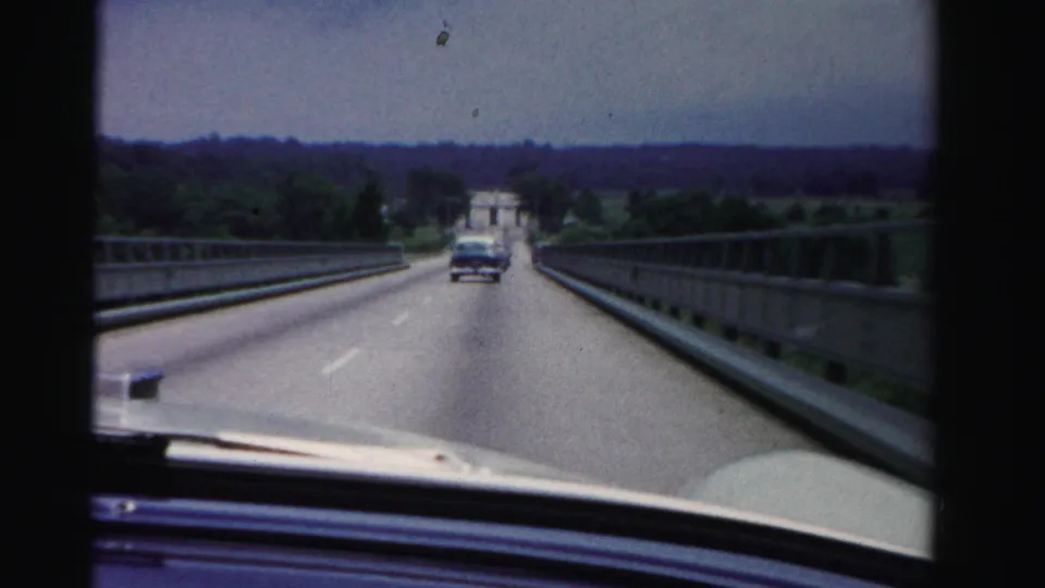 Forward road view from inside a moving car in Dade County, Florida, with a sedan ahead on a bridge and greenery in the distance.