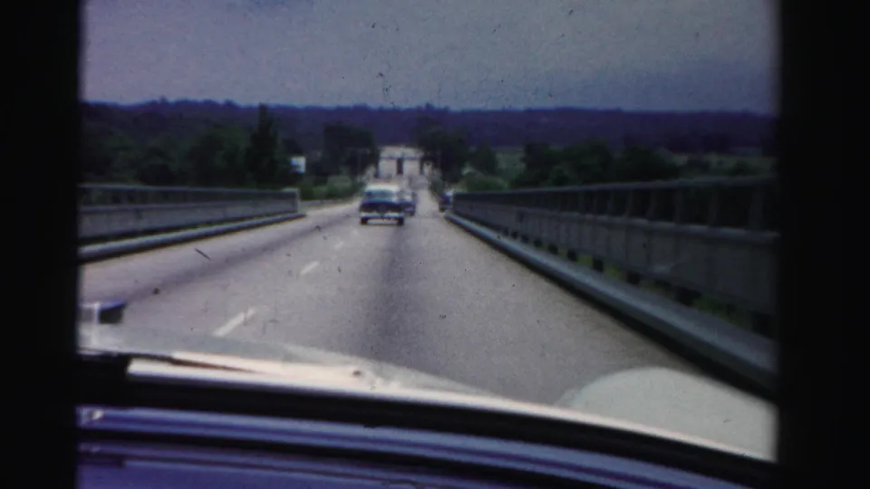 8mm home-movie frame of bridge driving in 1955 Dade County, Florida, with road markings, railings, dashboard edge, and a car ahead.