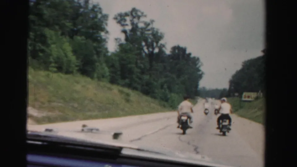 A 1955 home-movie frame shows motorcyclists ahead of the filming car on a broad Dade County roadway with wooded shoulders.