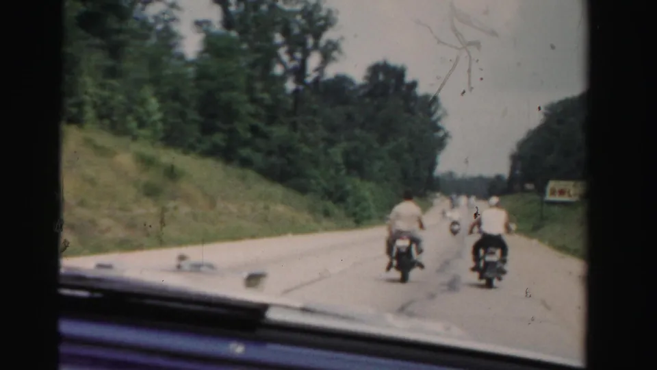 End frame from a 1955 Dade County driving shot, with motorcyclists continuing along a tree-lined road ahead of the camera car.