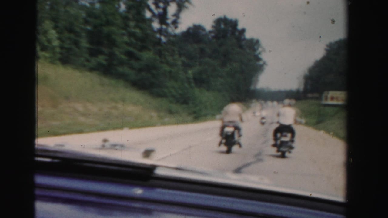1955 Dade County home-movie frame looking from inside a car toward two men on motorcycles traveling along a tree-lined highway.