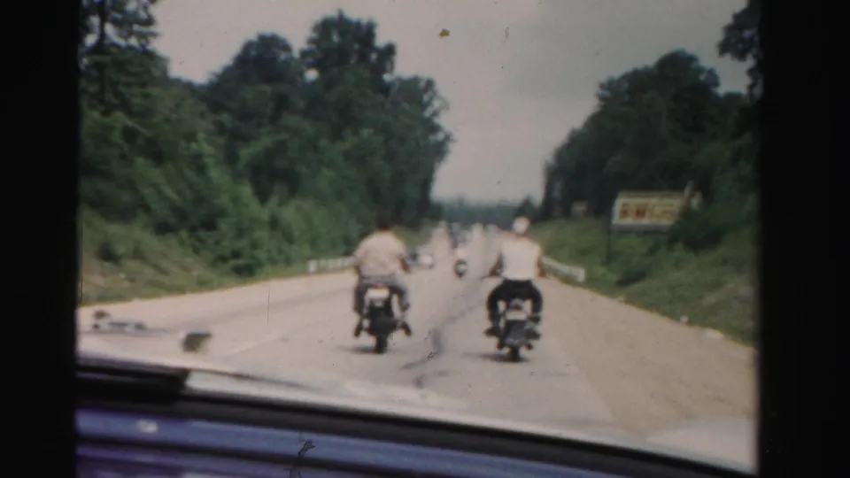 1955 Dade County 8mm frame from a moving car, with two nearby motorcycle riders and more riders farther ahead on a landscaped highway.