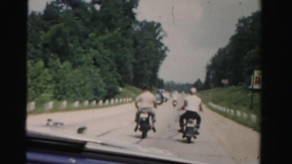 1955 Dade County home-movie frame showing motorcycle riders moving away along a gently curving highway with trees and roadside markers.