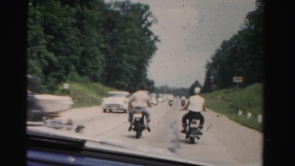 View through a windshield on a Dade County road, showing two motorcyclists, a passing car, roadside trees, and grassy shoulders.