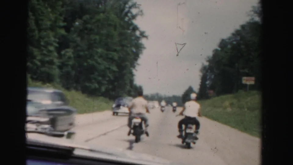 1950s Florida road scene with two motorcyclists ahead, a vintage car near the left lane, trees, roadside posts, and windshield framing.