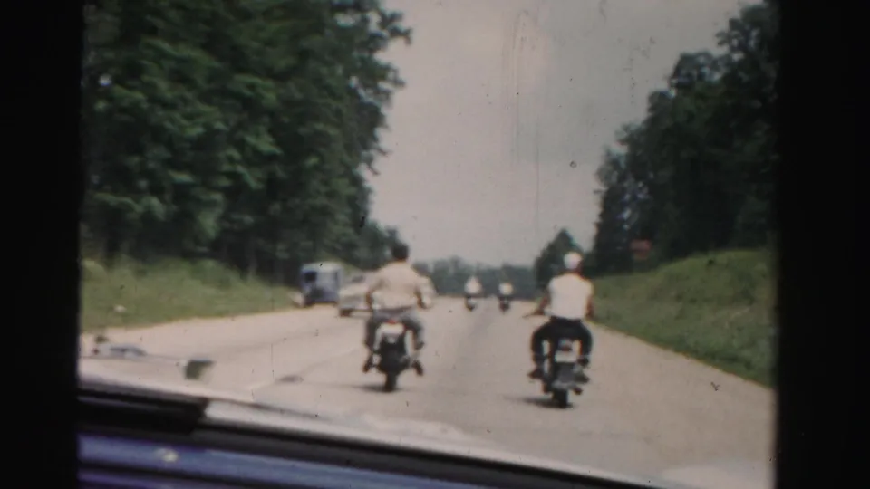 Dade County windshield view with motorcyclists ahead, distant vehicles, tree-lined shoulders, white markers, and soft 8mm film texture.