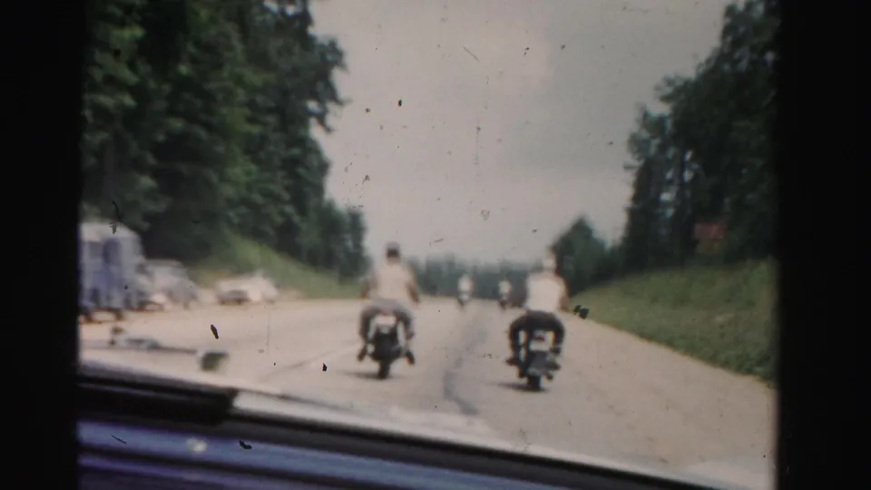 Two riders in light shirts continue along a Dade County road, framed by roadside trees, grassy banks, and distant mid-century traffic.