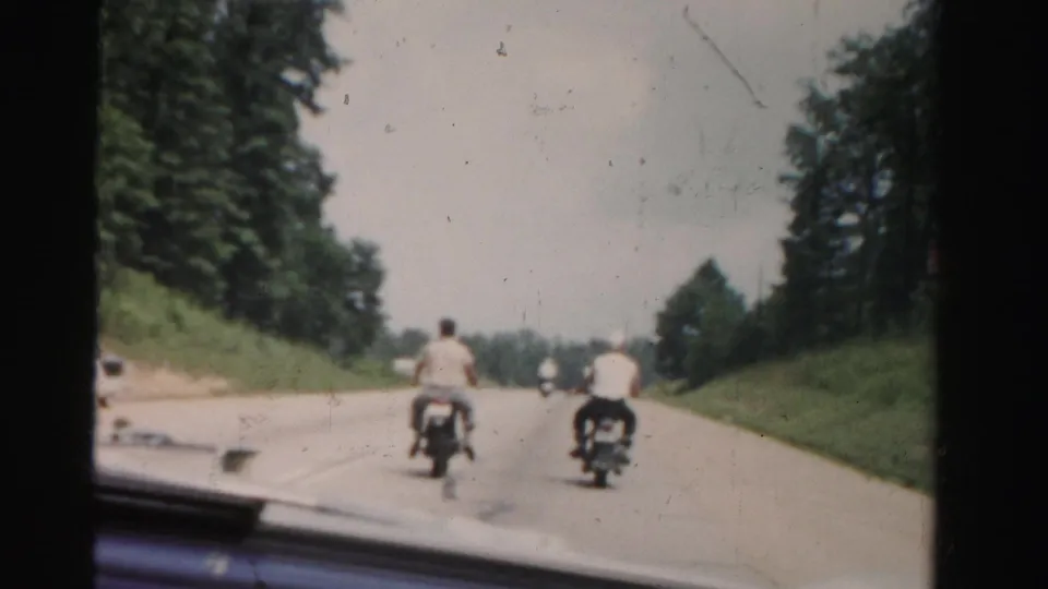 A soft 8mm frame shows two motorcycle riders moving away on a broad Florida road with dense trees and a grassy shoulder.