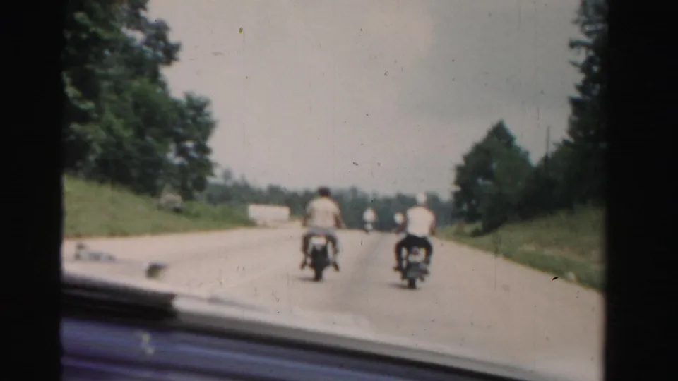 Two motorcycle riders remain ahead of the filming car on a tree-lined Dade County road, with distant vehicles and soft 8mm color fading.