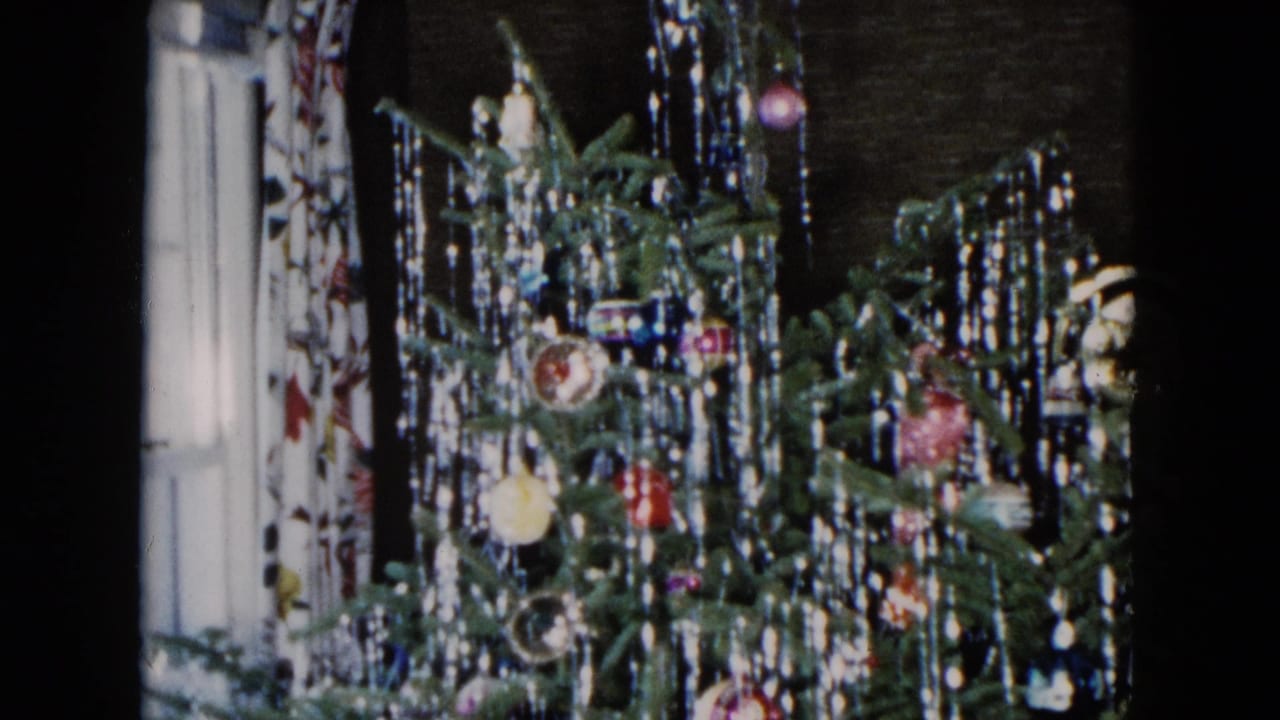 1955 Hicksville home-movie frame showing a Christmas tree with lametta tinsel, glass baubles, and a patterned curtain at left.