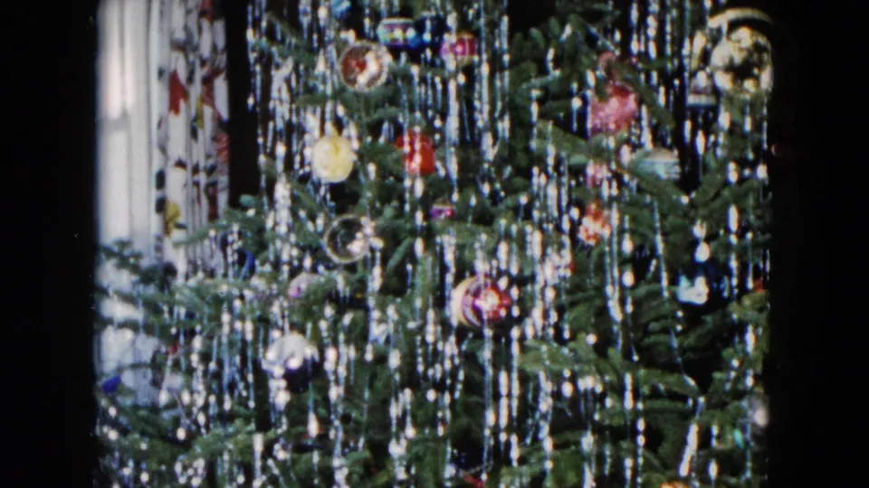 1955 Hicksville home-movie frame showing the lower tree area with lametta, colored ornaments, and a patterned curtain nearby.