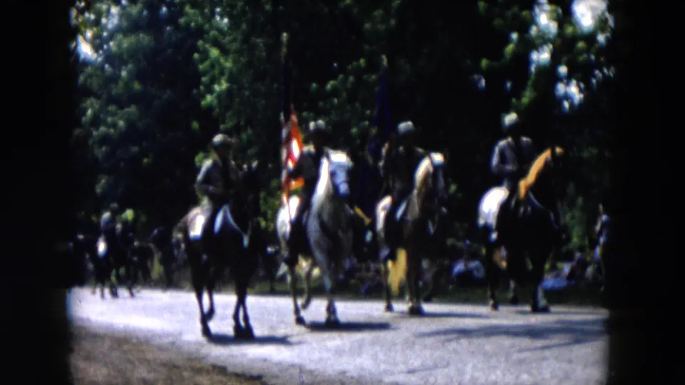 1955 Hicksville home-movie frame showing mounted parade riders, flags, trees, and roadside spectators from a shaded viewpoint.