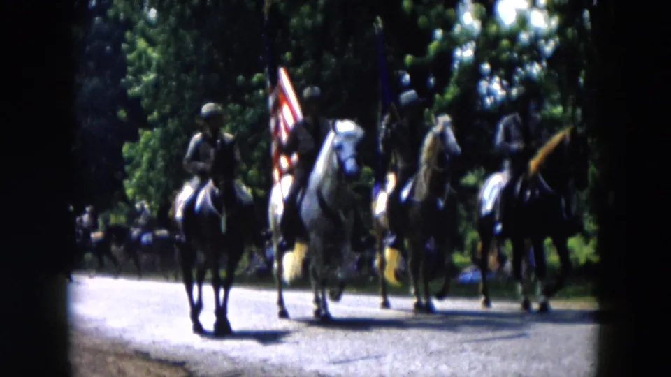 Mounted parade participants in Hicksville, New York ride past the camera with flags and spectators visible in the background.