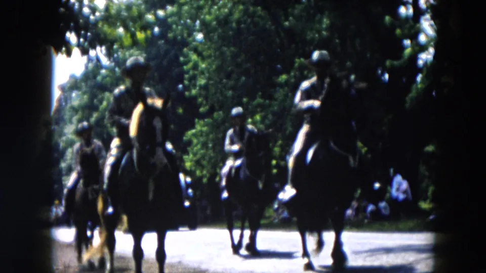 The mounted procession continues down the road in Hicksville, with spectators gathered near the trees in the background.