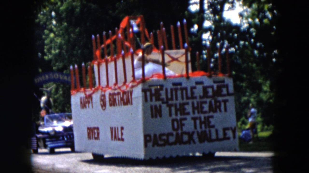 1955 home-movie frame of a River Vale birthday-cake parade float with red candles, Pascack Valley text, a dark car, and trees.