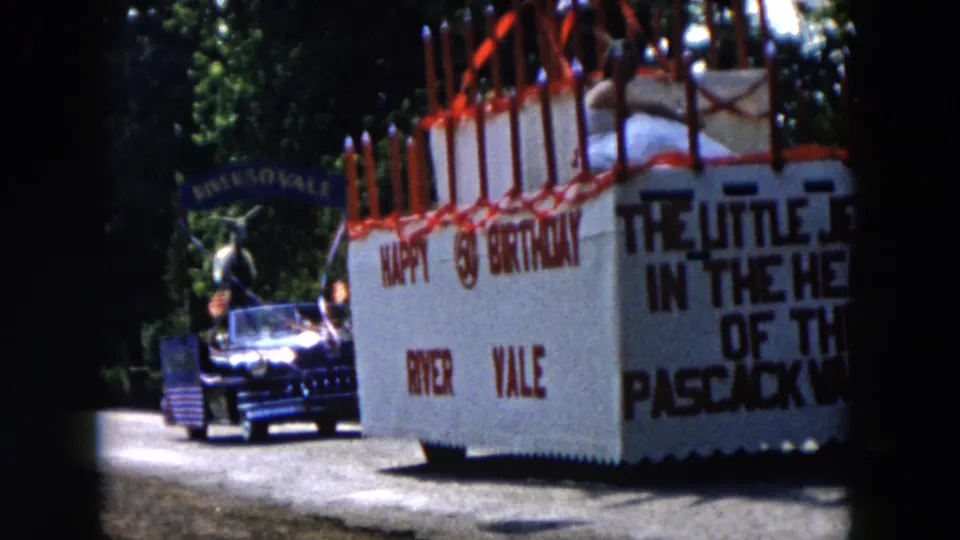River Vale birthday-cake float fills the frame, with red candles, slogan lettering, a dark car, and shaded trees visible.