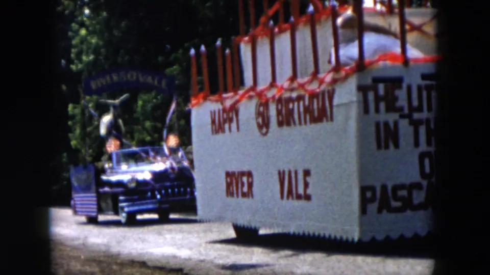 Close 1950s parade view of the River Vale birthday-cake float, showing red candle decorations, block lettering, and a following car.