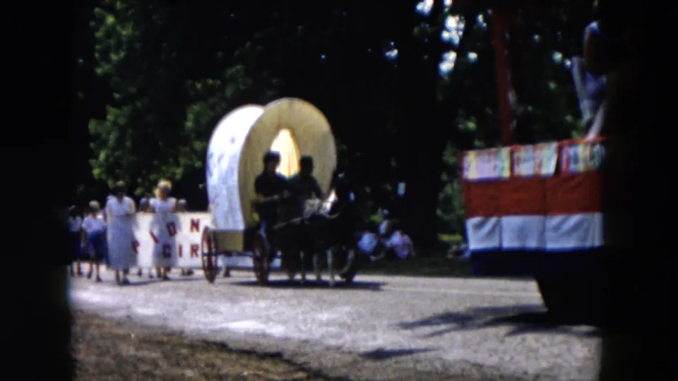A horse-drawn buggy with passengers appears on the parade route, with spectators, trees, and part of a red-and-white float nearby.