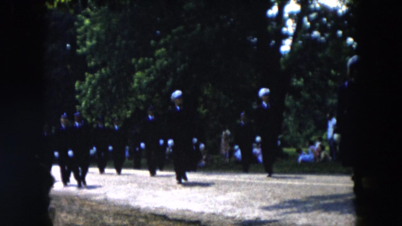 1955 Hicksville parade frame with dark-uniformed marchers wearing light gloves moving through a sunlit park setting.
