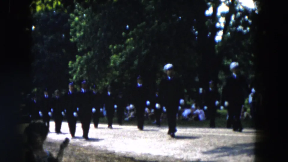 1955 Hicksville home-movie frame showing uniformed marchers passing spectators seated near trees during a parade.