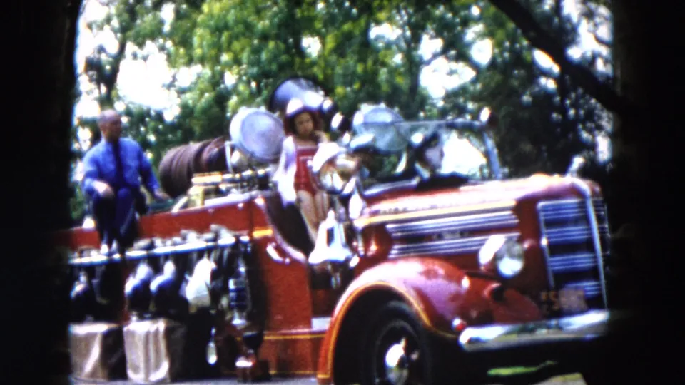 1955 Hicksville parade frame showing a red fire truck with chrome details and passengers passing under trees.