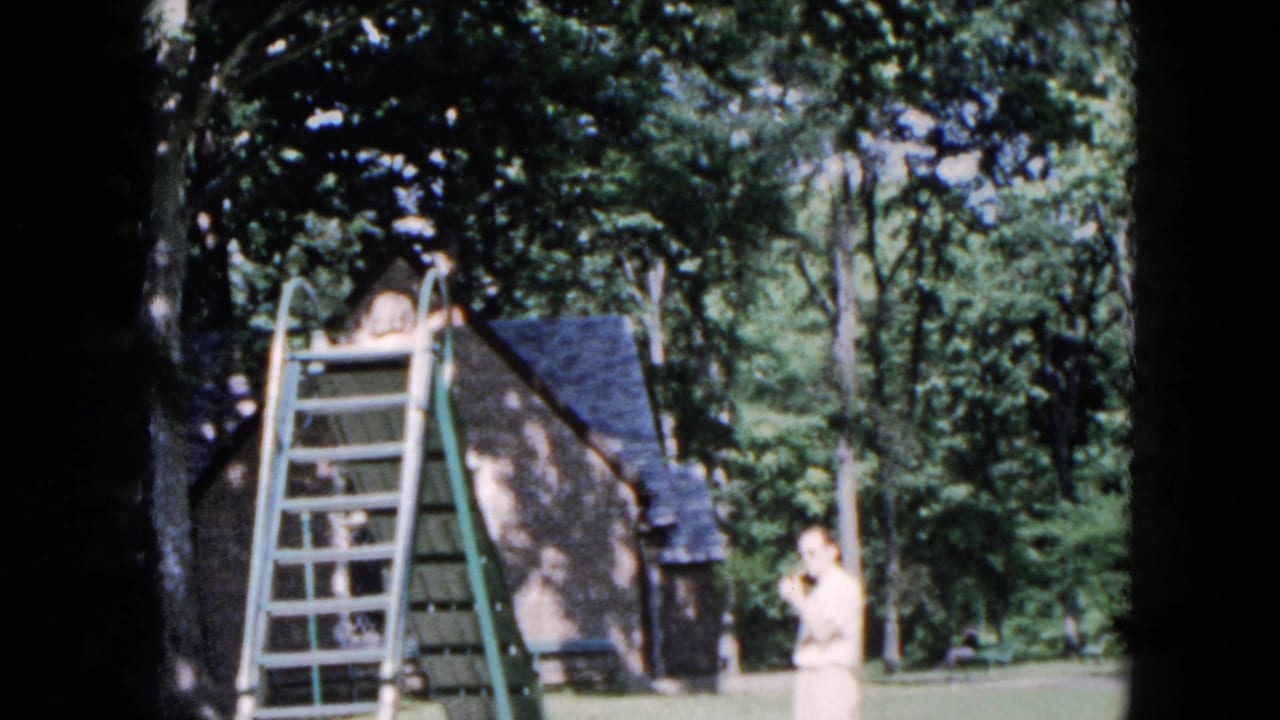 Boy climbs a green playground ladder while an adult stands nearby on a grassy lawn.