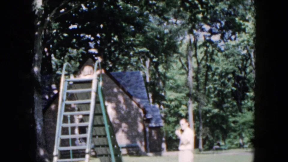 In 1955 Hicksville home-movie footage, a boy climbs a green metal playground ladder as an adult watches near trees and a small building.