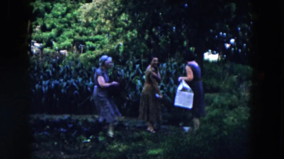 In the Hicksville home movie, three women in period dresses and hats gather in a garden, one holding a light-colored basket.