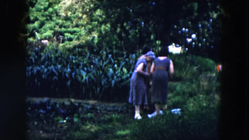 Two women in dresses stand close beside tall leafy plants in the 1955 Hicksville garden footage.