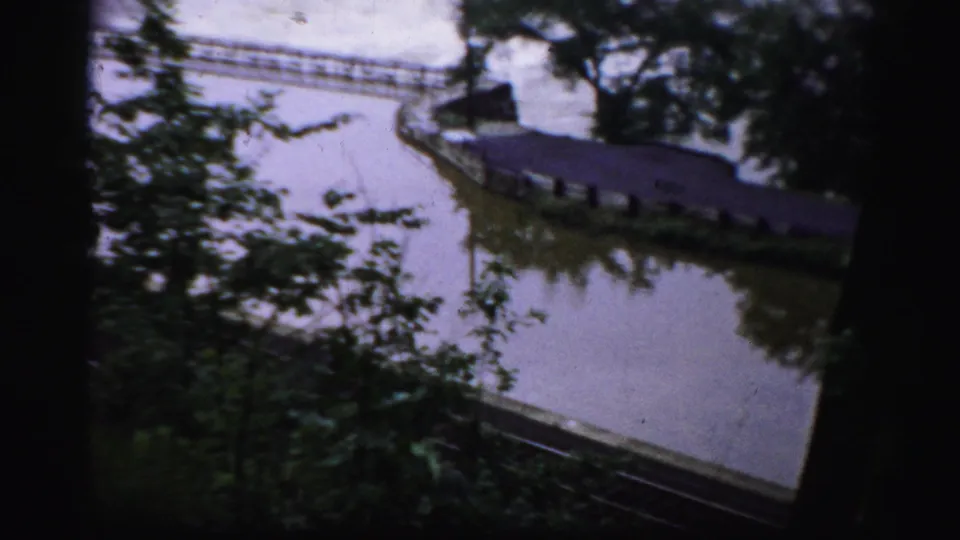 1950s Albany home-movie frame showing a riverbank, trees, a walkway or rail line, and water near the dam area.