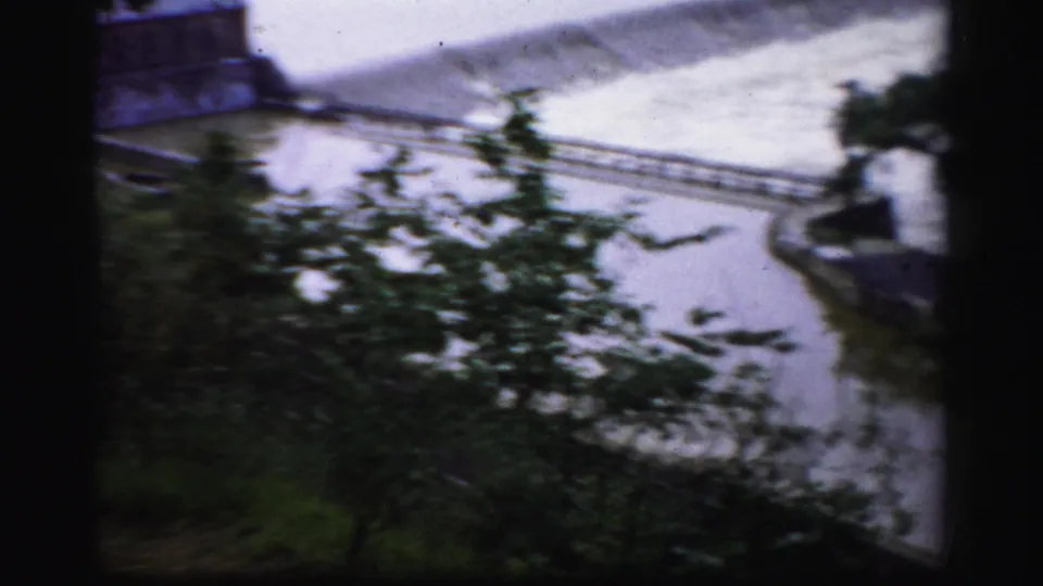 1950s Albany frame looking through foliage toward the dam spillway and a narrow walkway or rail structure over the water.