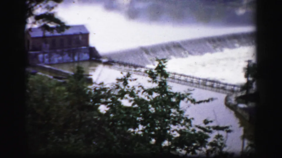 1950s Albany home-movie view of a mill-like building beside a dam spillway, partly obscured by foreground trees.