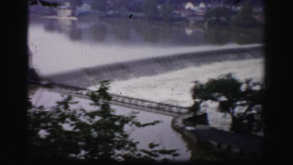1950s Albany frame showing the dam spillway, river water, foreground trees, and a low riverside building at the lower edge.