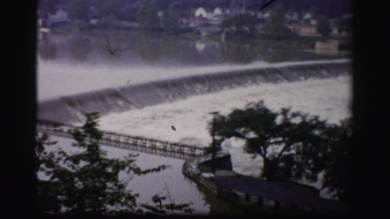Rushing river water spilling over a low dam with trees and a walkway in the foreground.