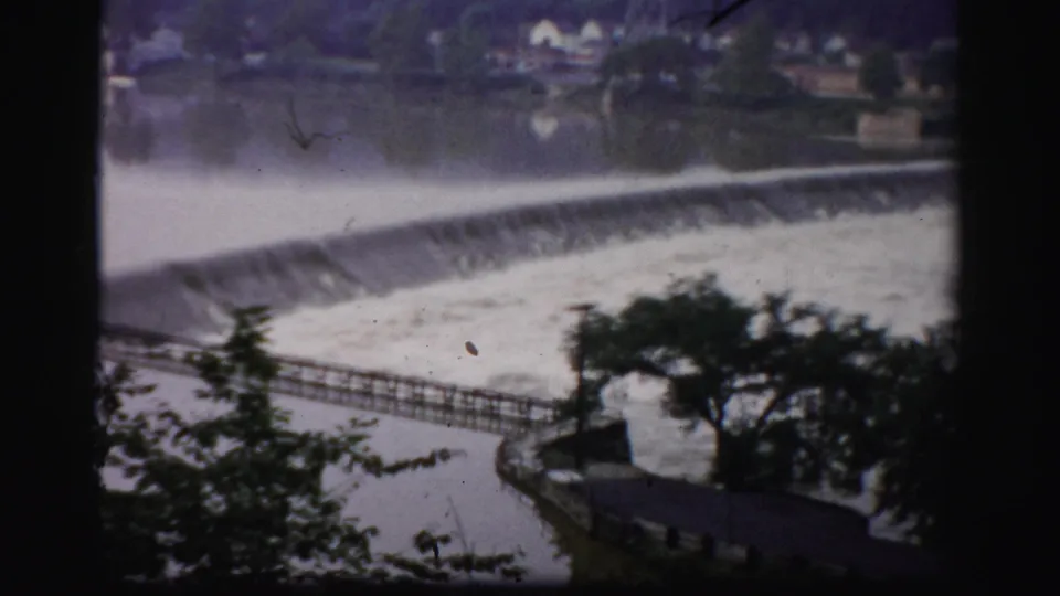1950s Albany-area home-movie view of a river pouring over a low dam, framed by trees and a riverside walkway.