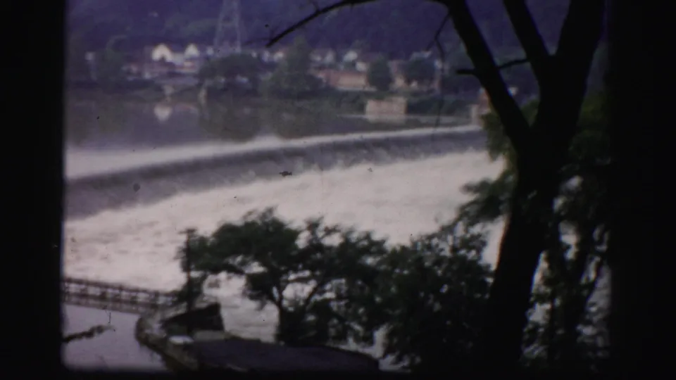 Color 8mm frame of an Albany-area river spillway with white water, railings, trees, and distant buildings.