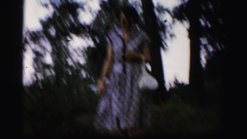 Brief 1950s home-movie glimpse of a woman in a patterned dress walking through trees near the riverside.