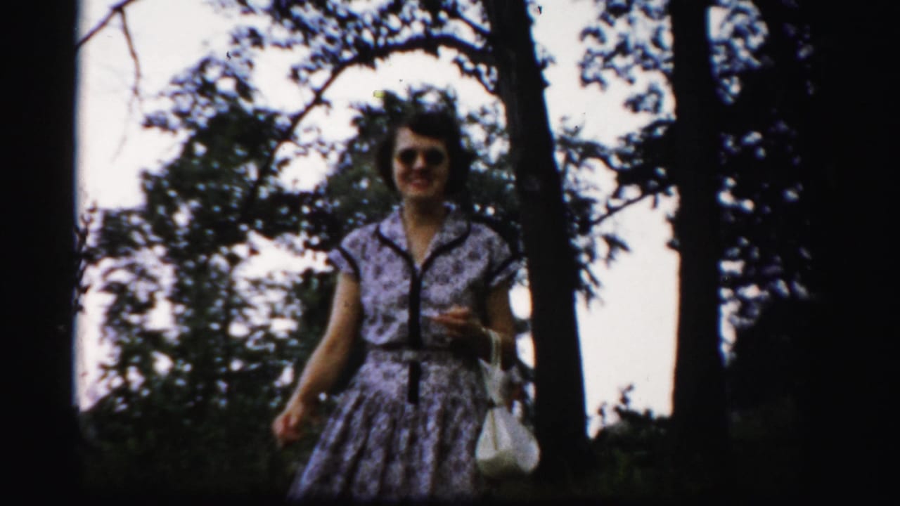 In 1956 Albany home-movie footage, a woman in sunglasses and a floral dress walks through a wooded lakeside area.