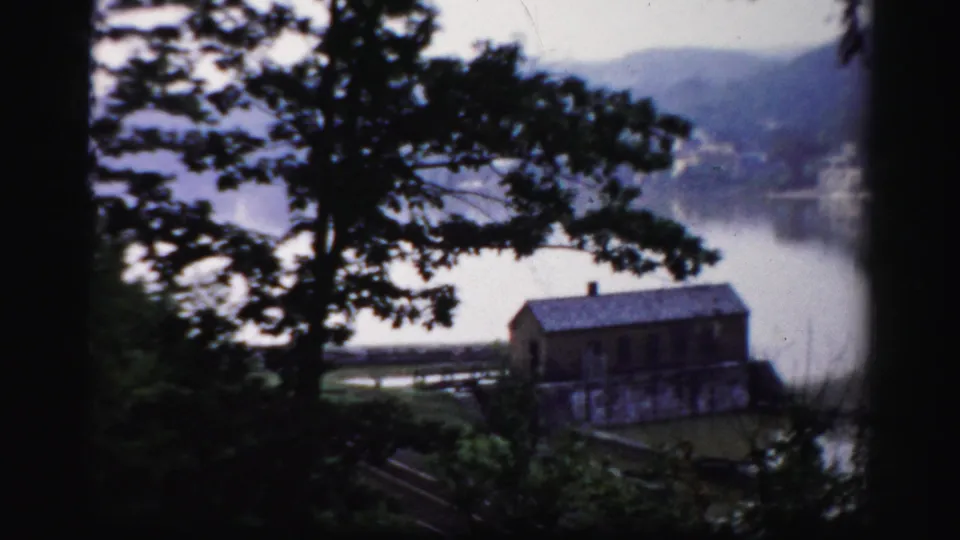 The camera looks through trees toward a lakeside building, water, and hazy hills in 1956 Albany home-movie footage.
