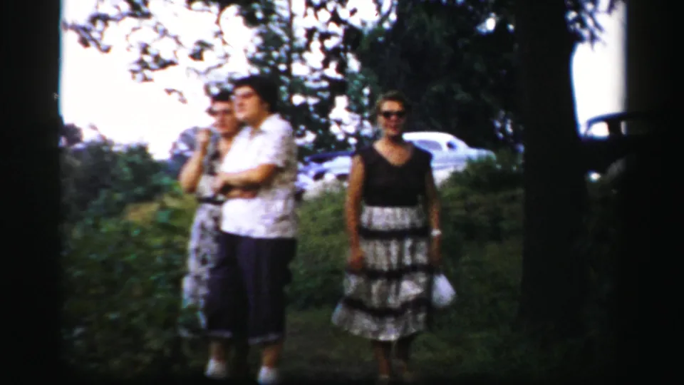 Three adults stand in a grassy Albany yard in 1950s home-movie footage, framed by trees and parked cars.