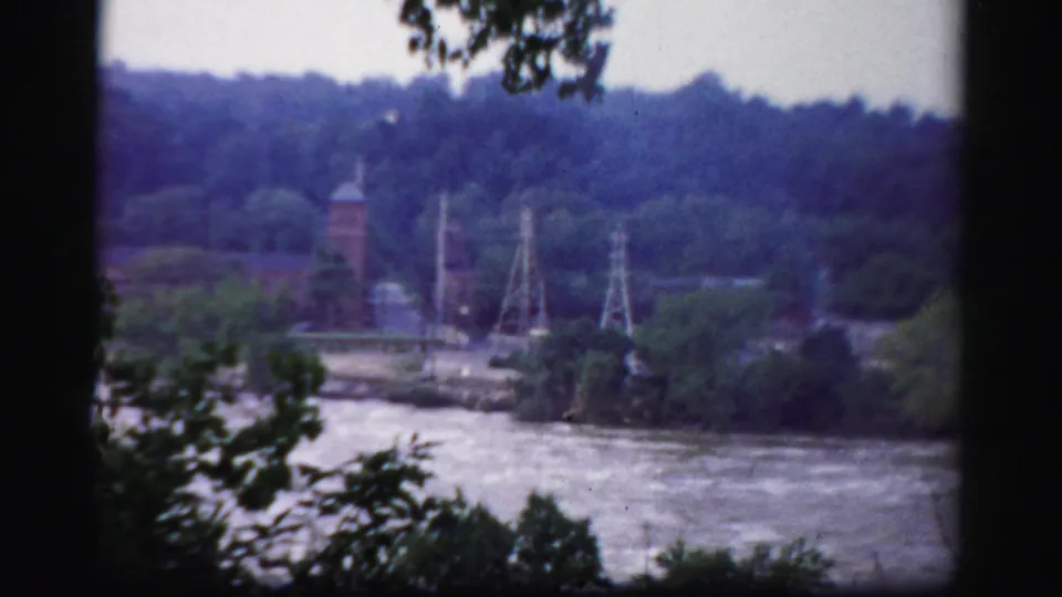 The camera opens to a riverside view near Albany, showing water, trees, wooded hills, utility structures, and a distant tower.