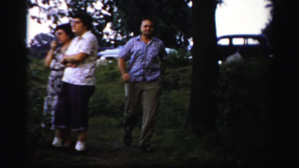 Three adults in casual clothing stand near trees in Albany-area home-movie footage, with a vintage car behind them.