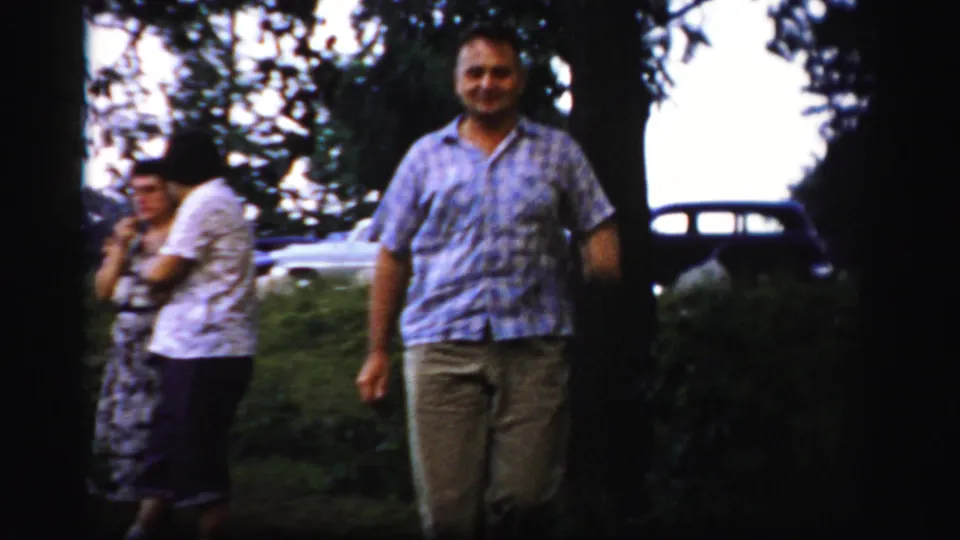 An adult man in a checked shirt walks toward the camera while two adults and a parked car remain in the background.