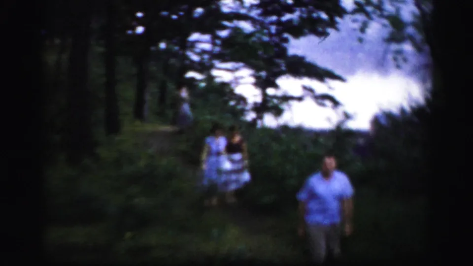People approach along a green riverside path, with dark trees and bright water in the background in 1950s Albany-area home-movie footage.